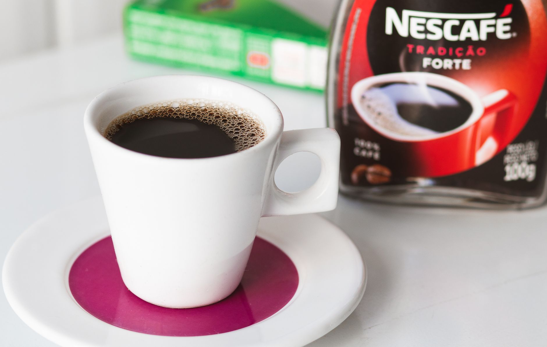Biały kubek czarnej kawy na różowym spodku, słoik Nescafé w tle